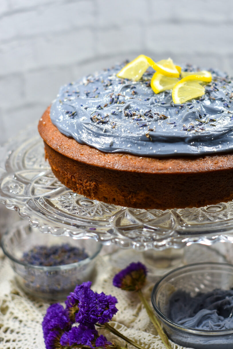 An Earl Grey frosted sponge cake on a crystal cake stand, surrounded by lace and purple flowers