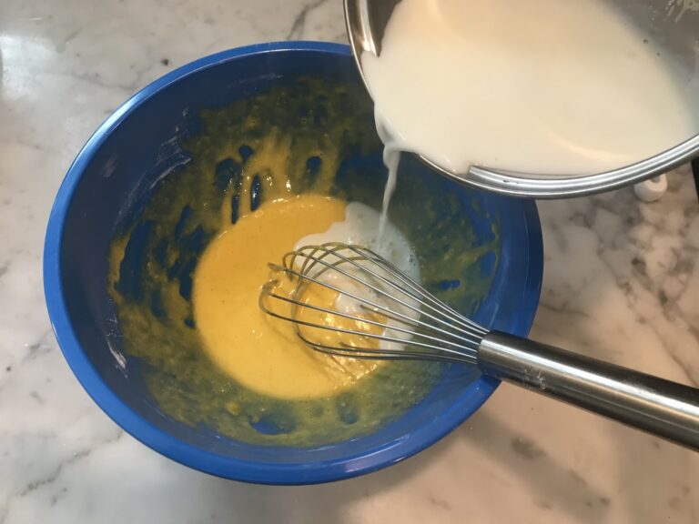 Tempering hot milk into a mixing bowl containing egg mixture and a metal whisk