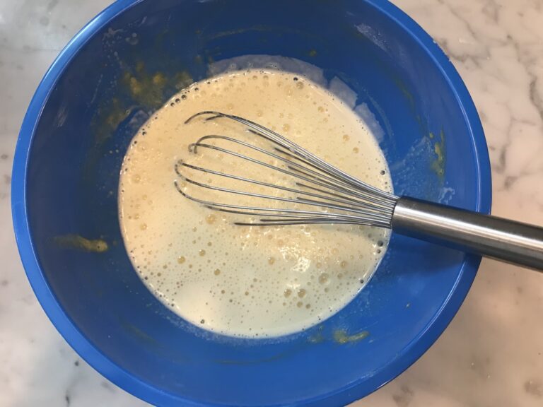 A blue mixing bowl filled with tempered pastry cream mixture, and a metal whisk