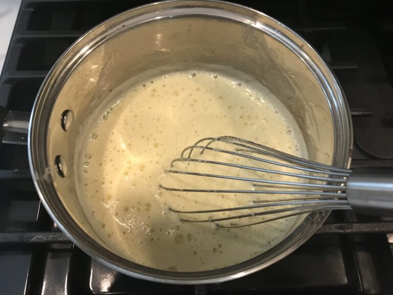 Metal saucepan on stovetop, containing tempered pastry cream mixture and a whisk