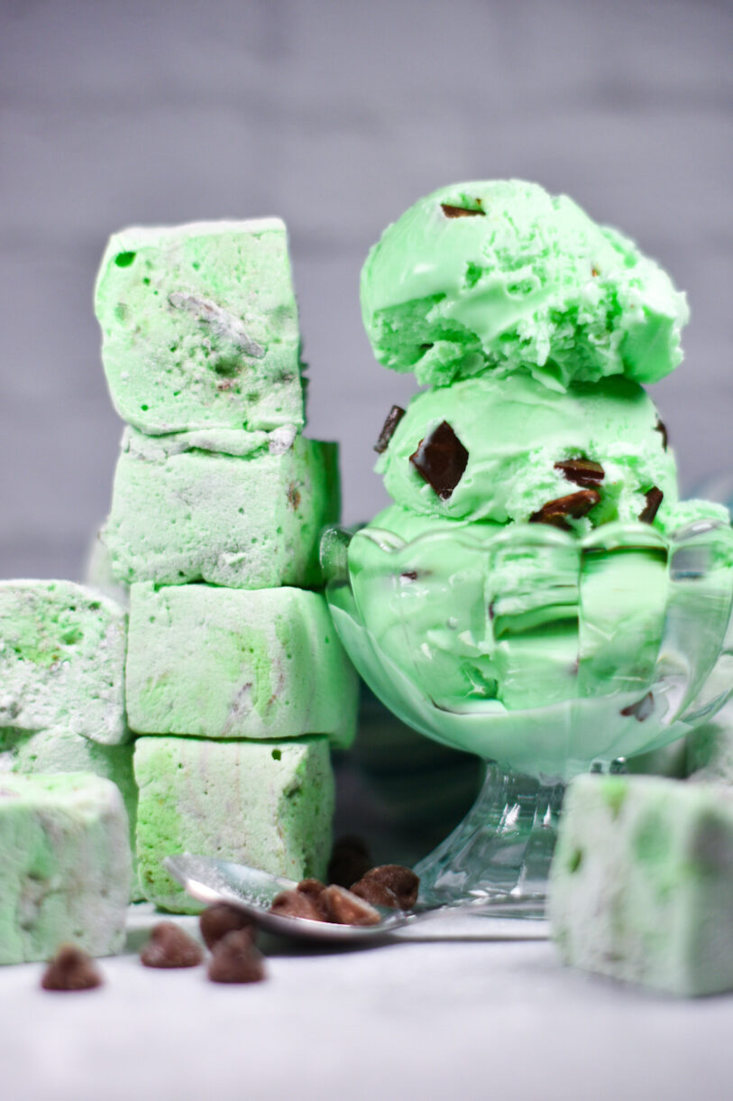 A bowl of mint chocolate chip ice cream next to a stack of mint green homemade marshmallows