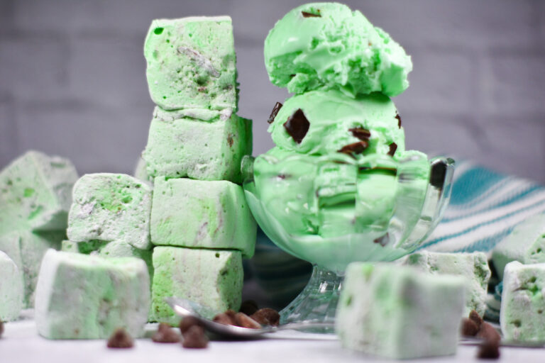 Horizontal shot of a glass bowl of mint chocolate chip ice cream next to a stack of mint chocolate chip marshmallows