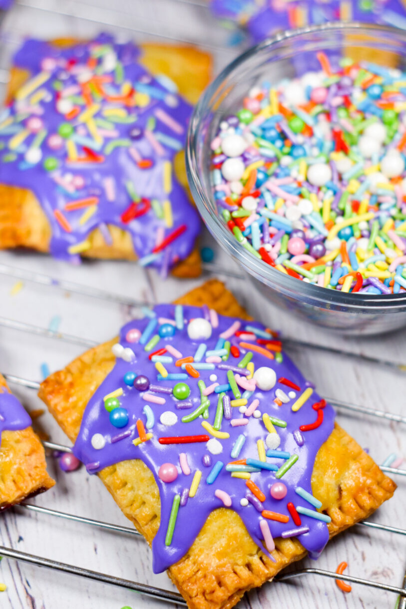 Frosted blueberry hand pies and a clear glass bowl of sprinkles on a white wooden surface