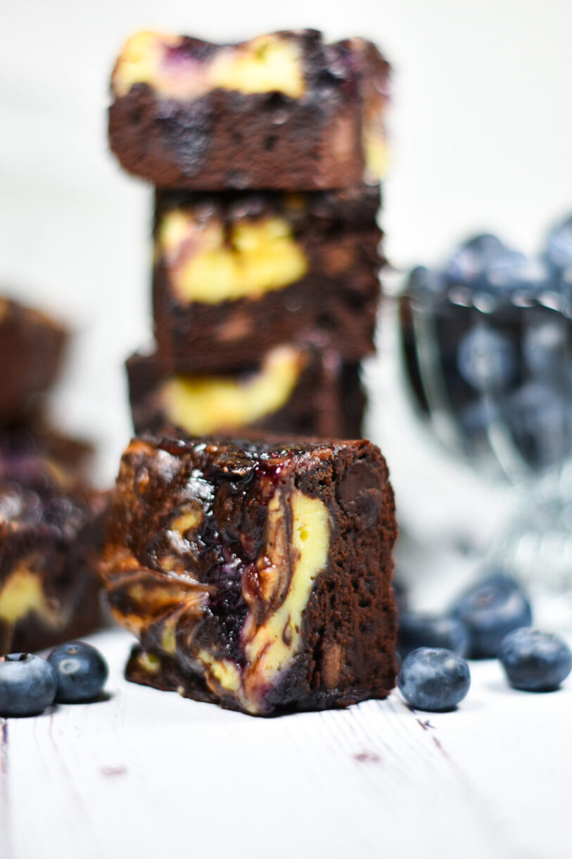 A blueberry cheesecake brownie surrounded by fresh blueberries, with a stack of brownies in the background