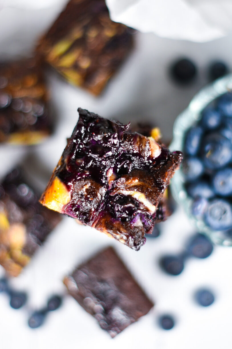 The top of a blueberry cheesecake brownie, with additional brownies and blueberries out of focus in the background
