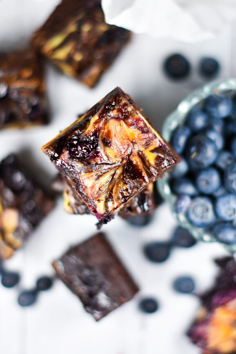 A stack of blueberry lemon cheesecake brownies, with a bowl of blueberries, on a white background