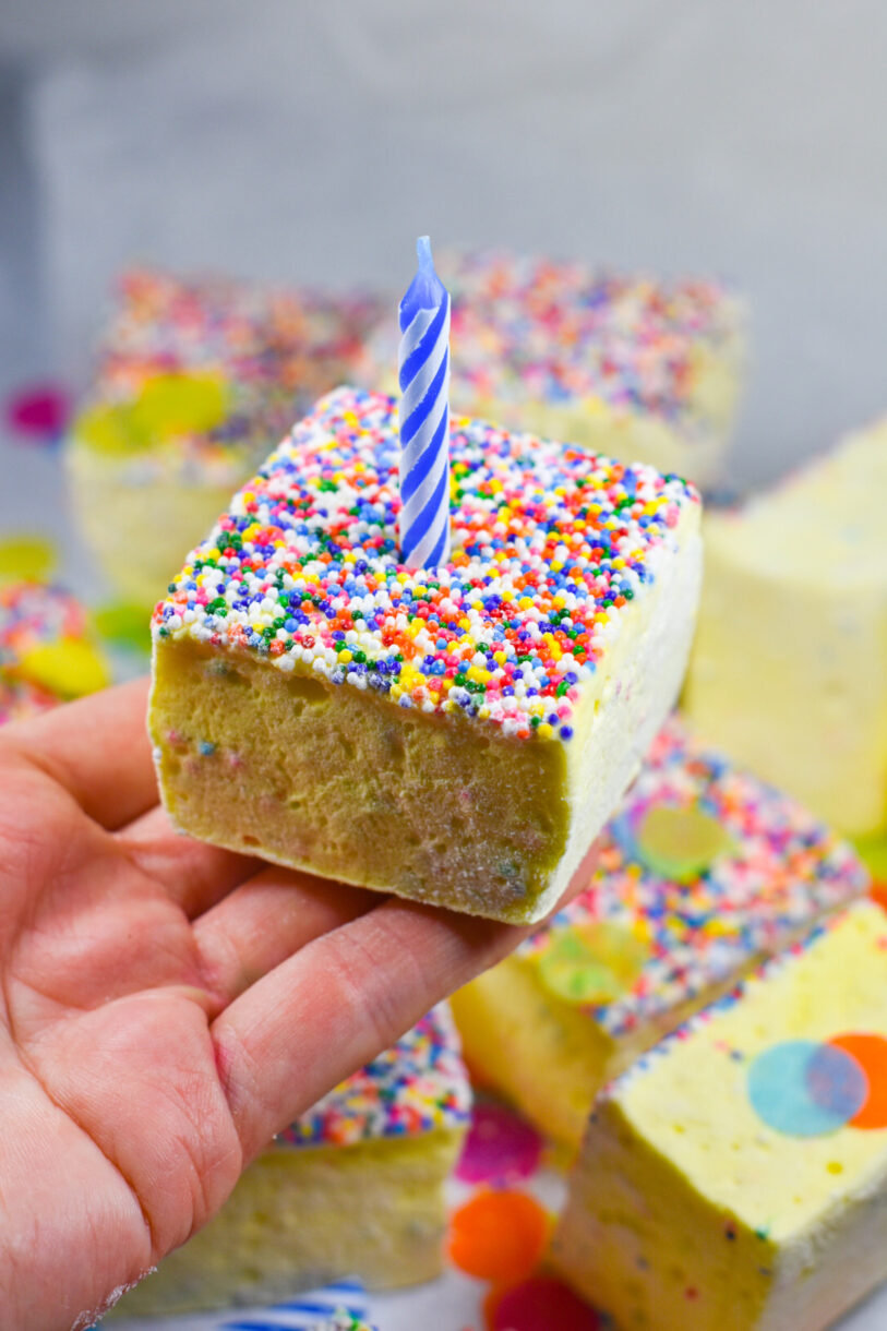 A hand holding a slice of birthday cake marshmallow with a blue and white candle on top