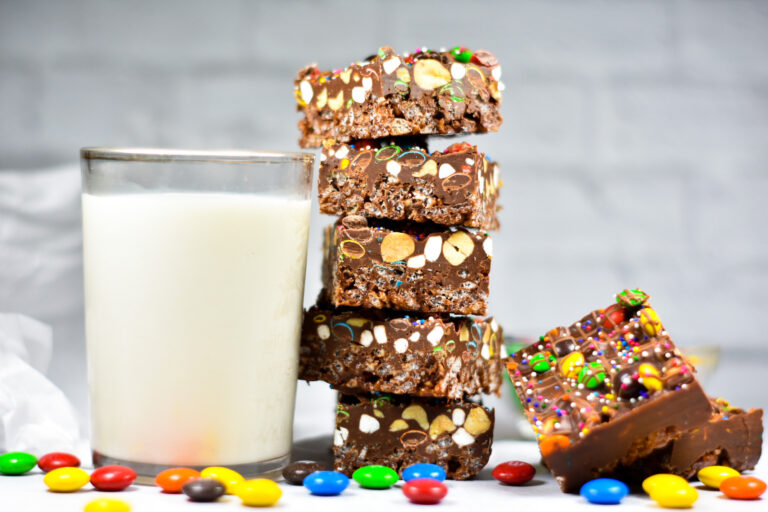 A stack of rainbow rocky road slices next to a glass of milk, with a white brick background behind
