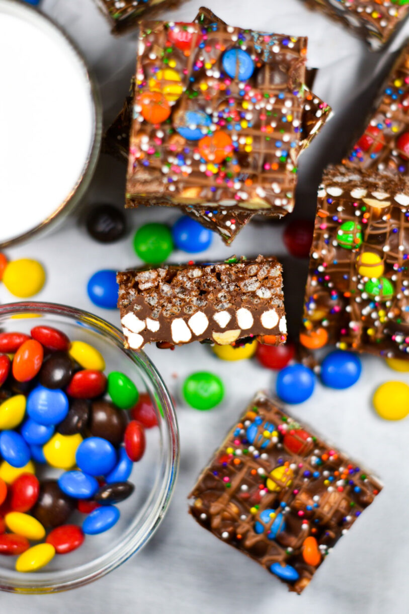 Rainbow rocky road slices, a bowl of M&Ms, and a glass of milk, on a white background
