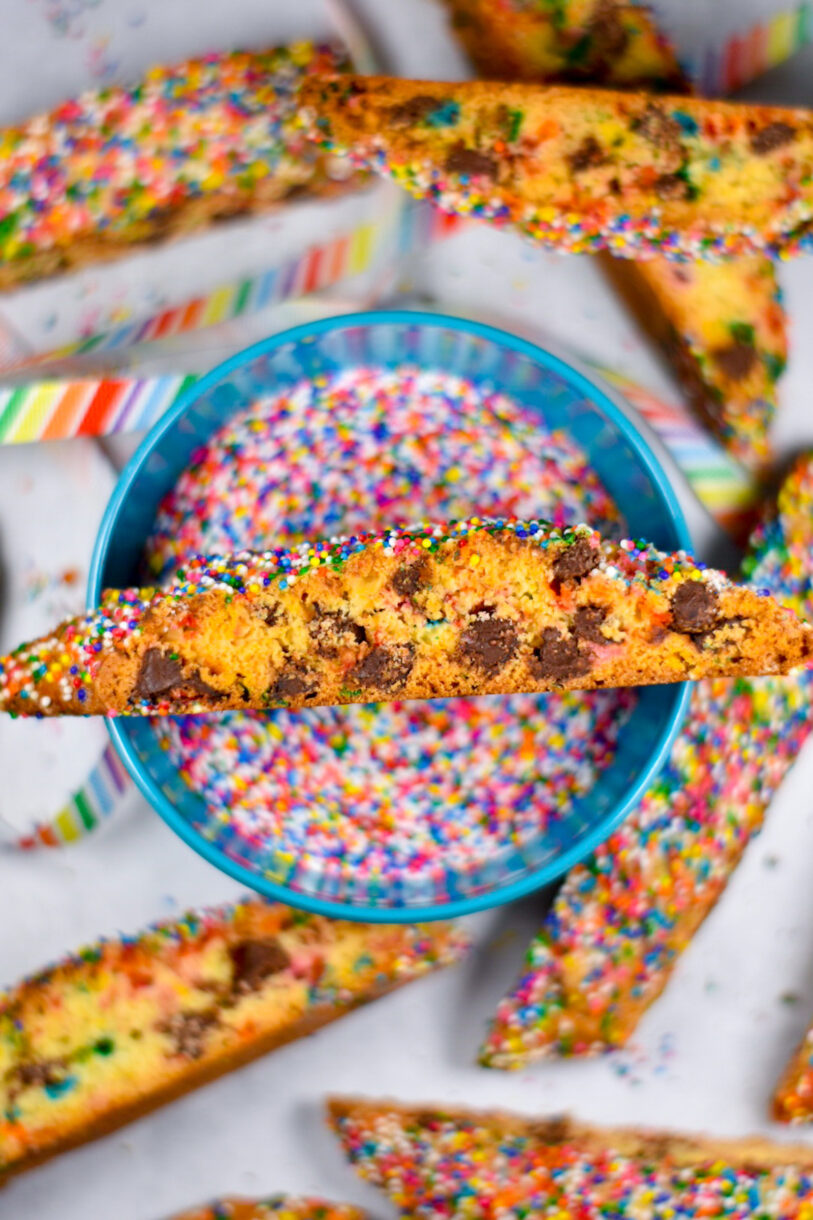 A bowl of rainbow sprinkles with a slice of chocolate chip cookie balanced on the top