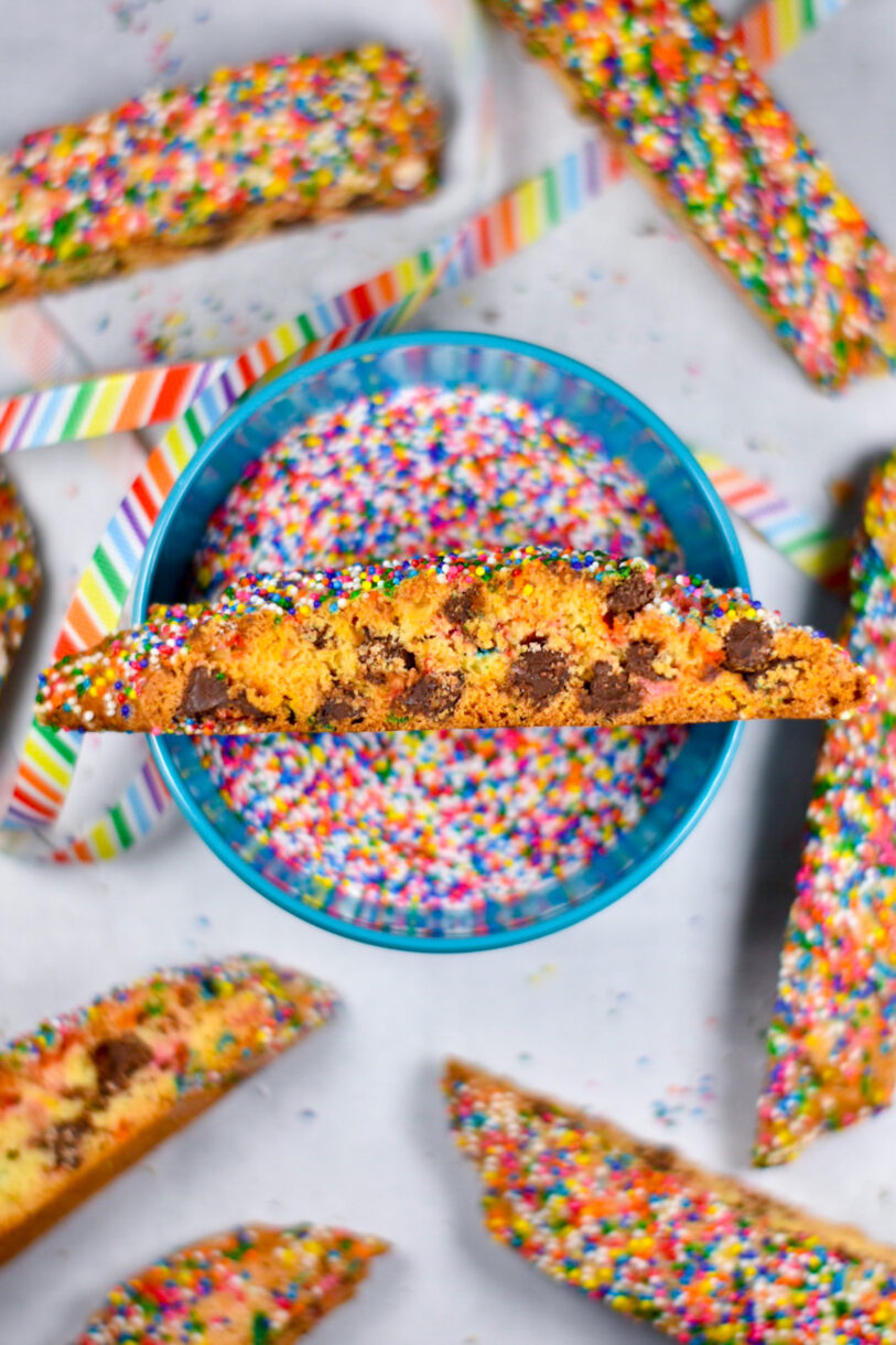 Rainbow ribbon, a blue bowl of rainbow sprinkles, and a slice of biscotti on the top