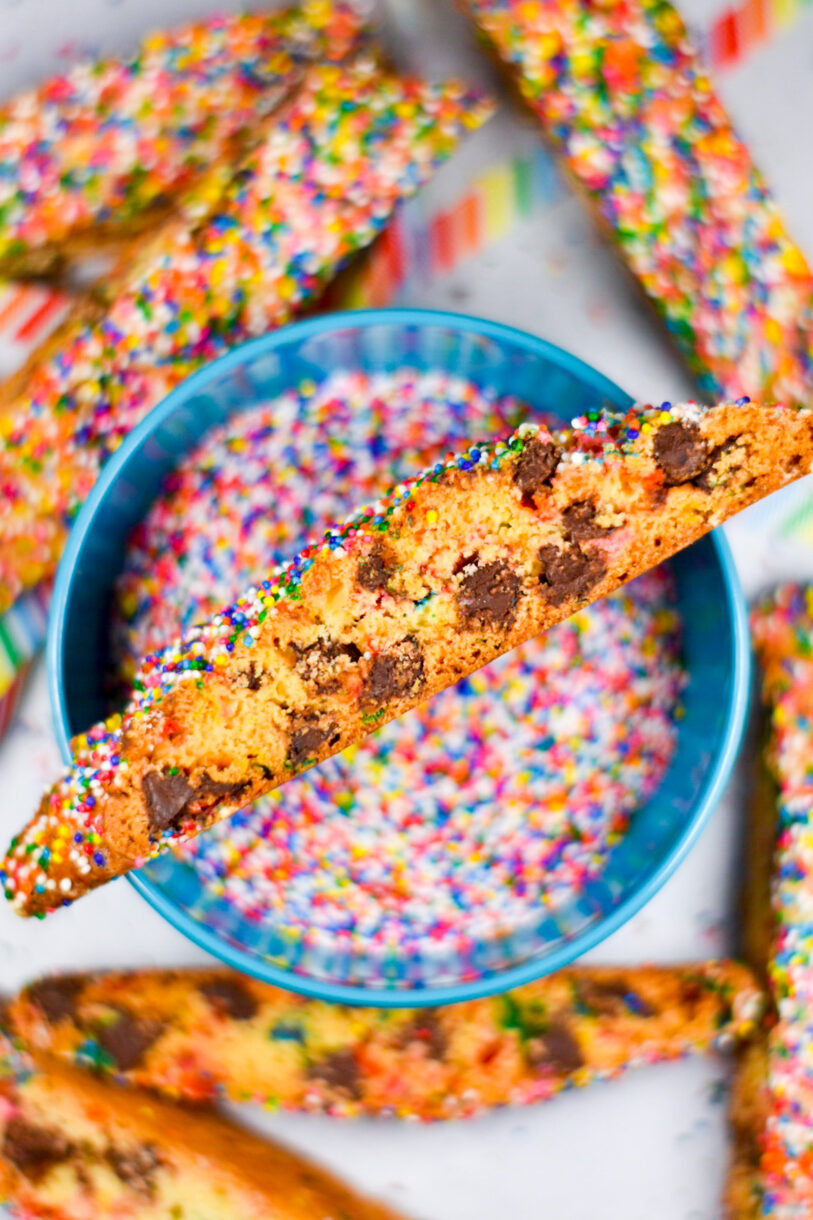 Chocolate Chip Rainbow Biscotti sitting on a bowl of rainbow nonpareil sprinkles