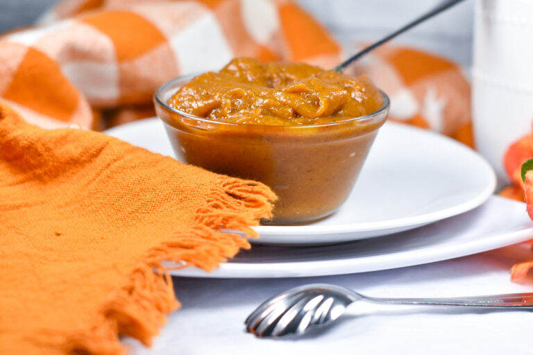 A glass bowl of pumpkin spread with orange tea towels, white plates, and a metal spoon