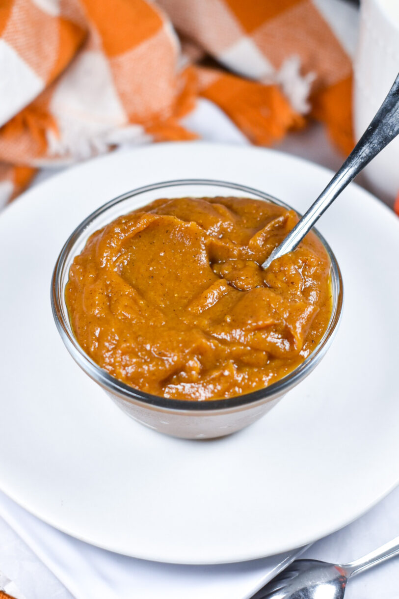 A glass bowl of spiced pumpkin butter sitting on a white plate, with a metal spoon