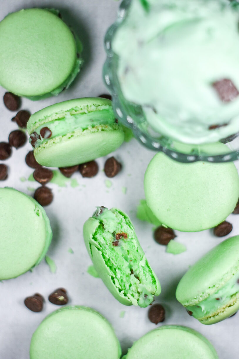 A bowl of mint chocolate chip ice cream, macarons filled with buttercream, and chocolate chips on a white background