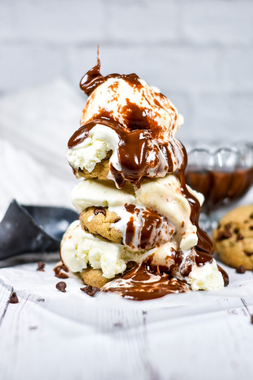 Homemade hot fudge sauce drizzled on a stack of cookies and ice cream, with an ice cream scoop and bowl of fudge sauce in the background