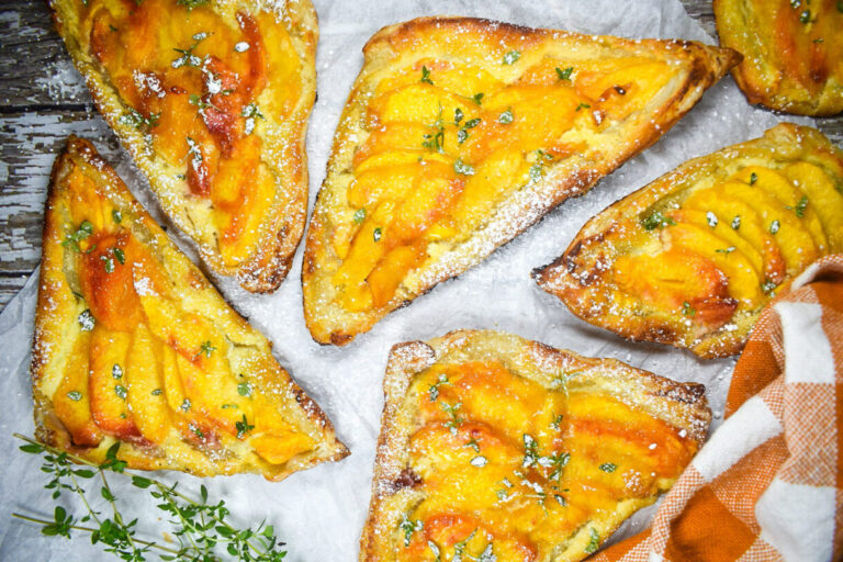 Peach pastries arranged on a wooden surface with a sheet of white parchment, gingham tea towel, and thyme