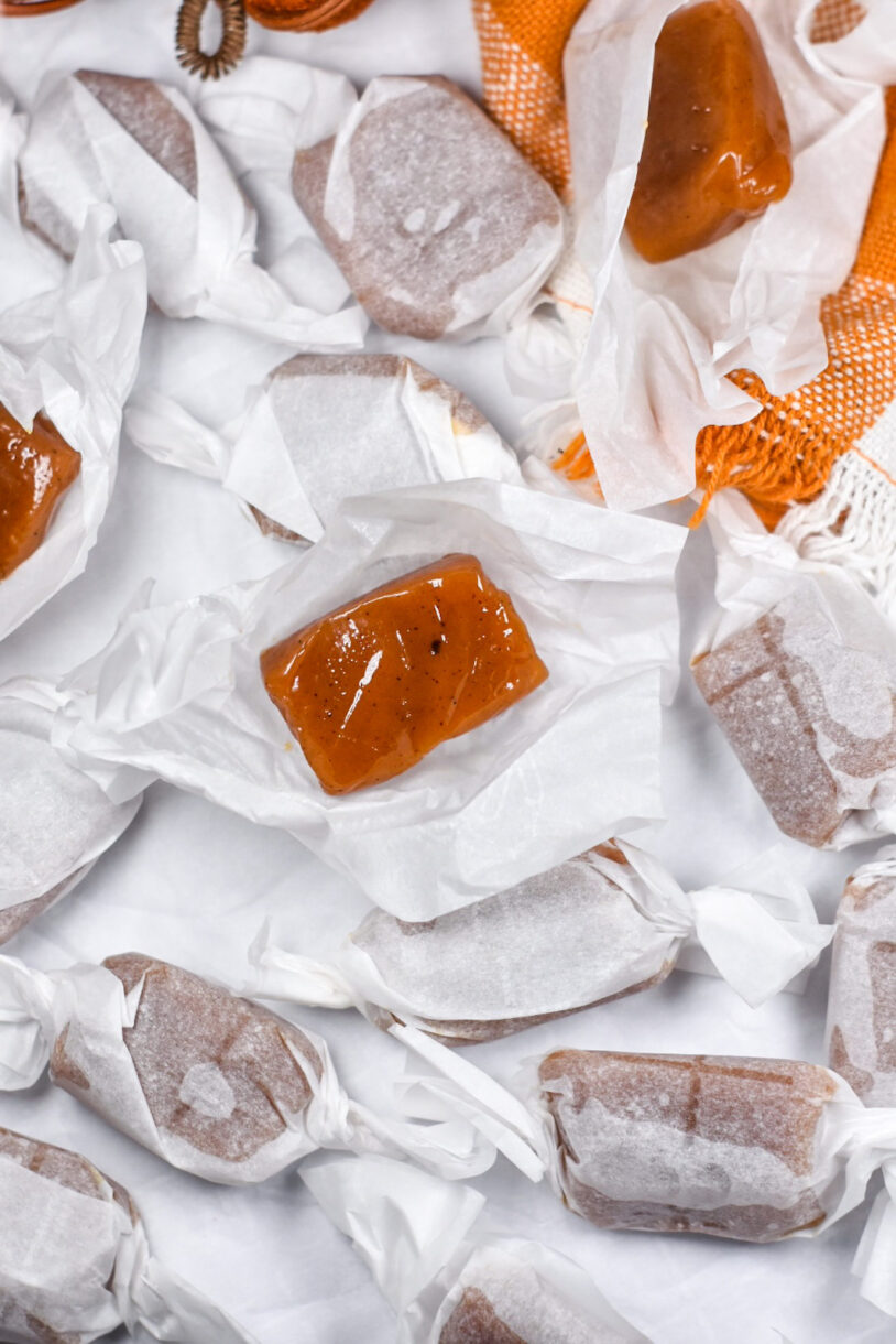 Vertical shot of a pair of unwrapped caramels on squares of parchment, surrounded by wrapped pumpkin caramels