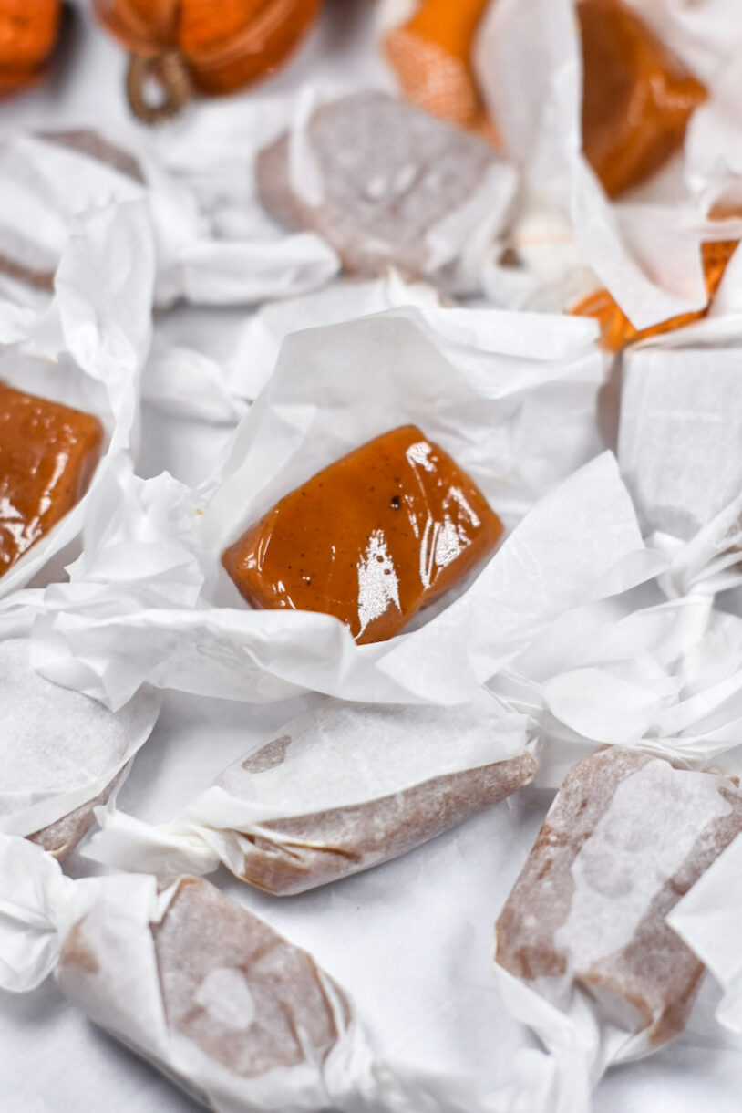 A pair of unwrapped caramels on squares of parchment, surrounded by wrapped pumpkin caramels