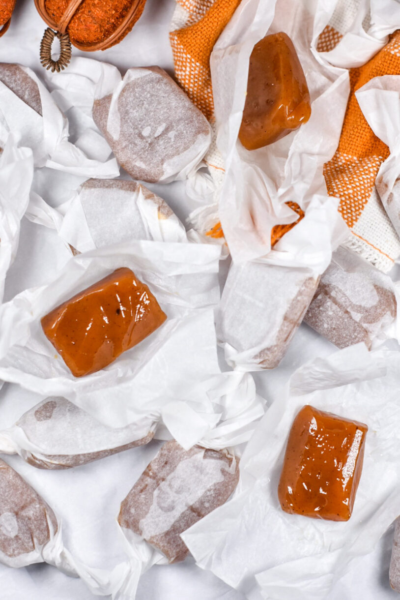 A pair of unwrapped caramels on squares of parchment, surrounded by wrapped pumpkin caramels