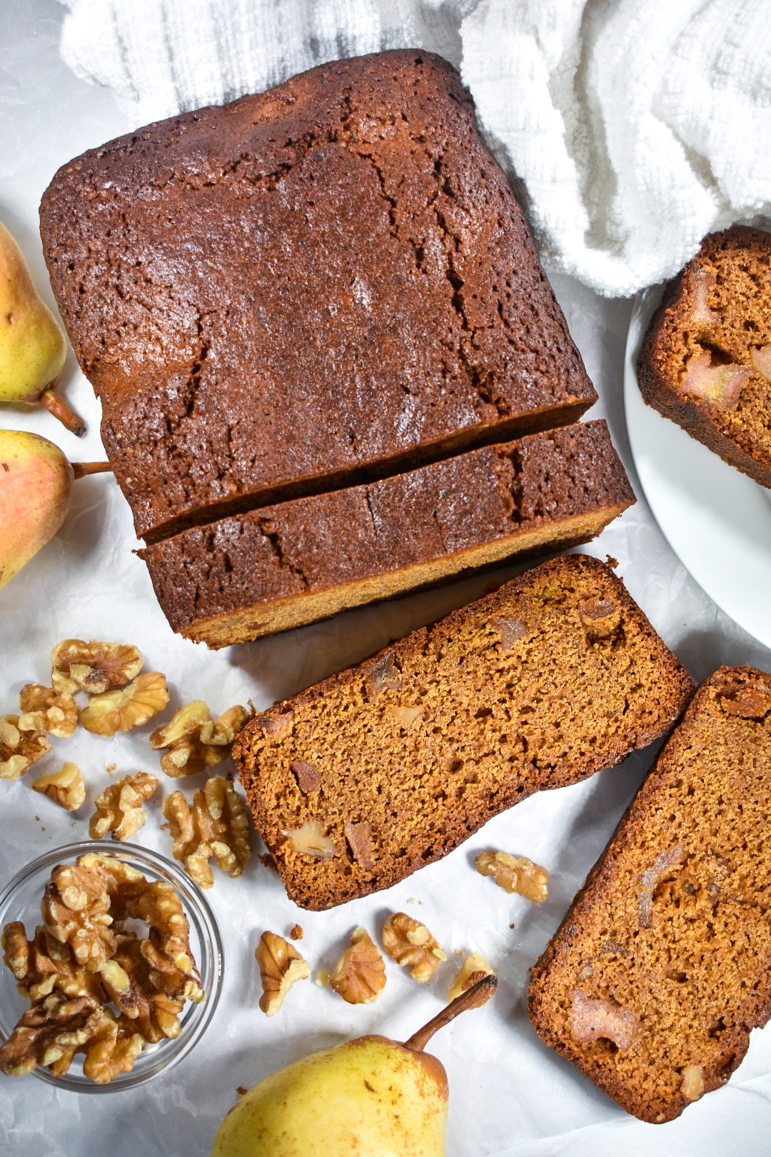 Gingerbread loaf with slices cut, on a white background with whole pears and a bowl of walnuts