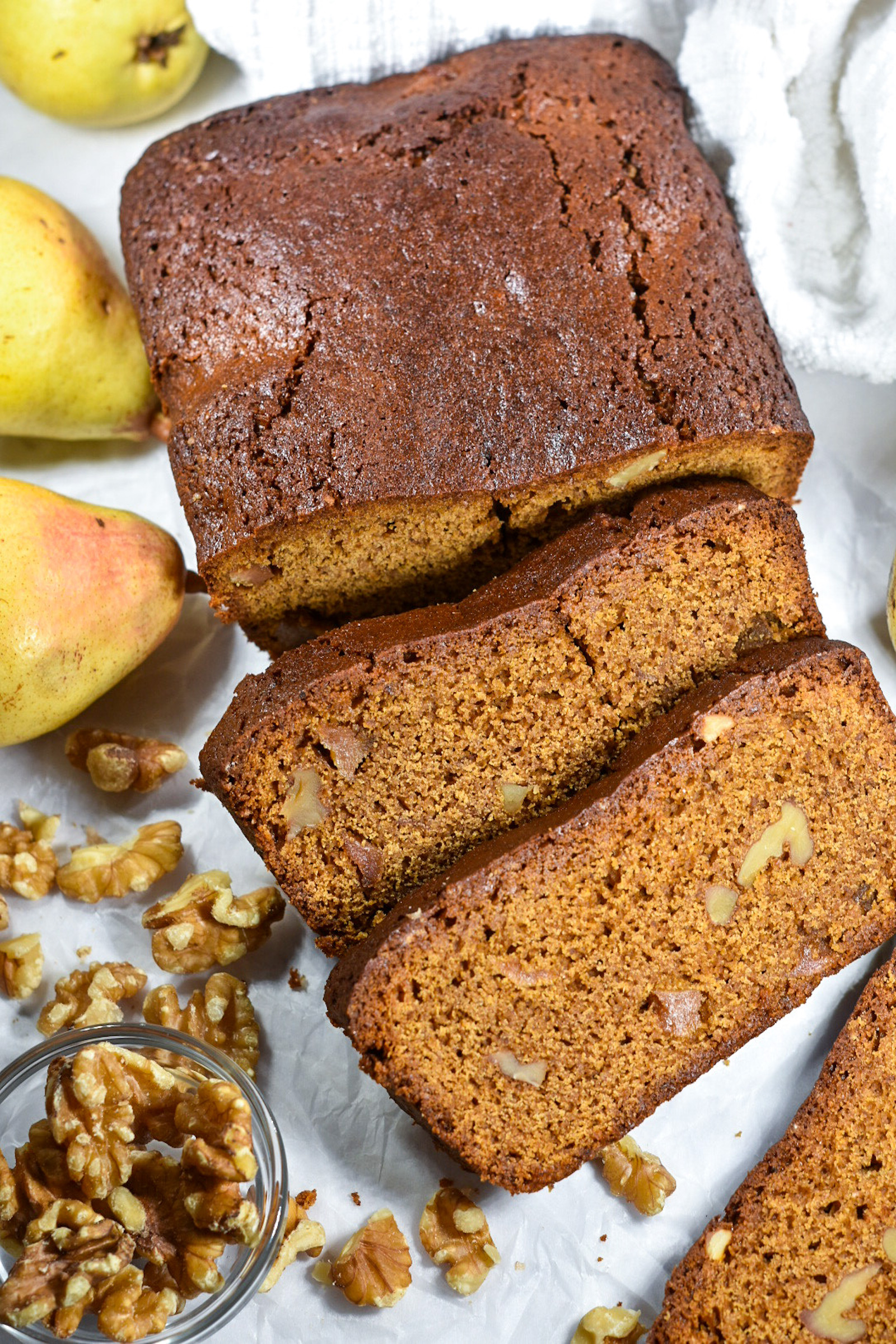 Gingerbread Loaf with Walnuts and Caramelised Pears, alongside whole pears and a bowl of walnuts, on a white background