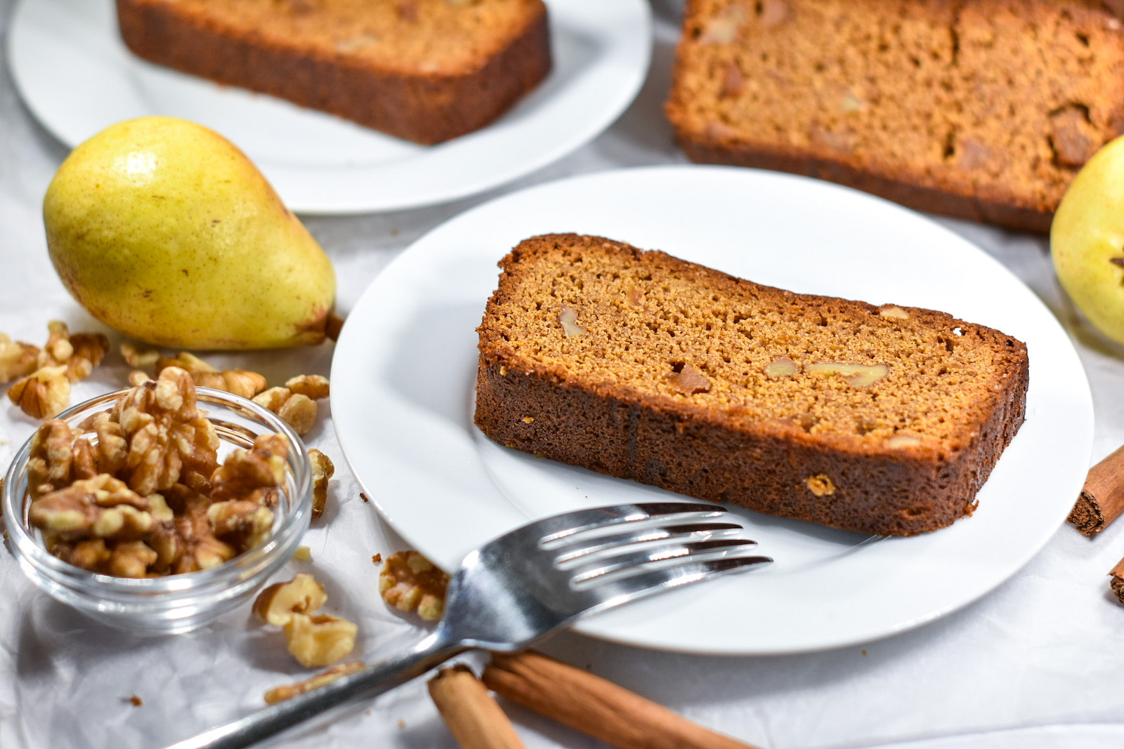 Gingerbread loaf slice on a plate, on a white background with whole pears and a bowl of walnuts
