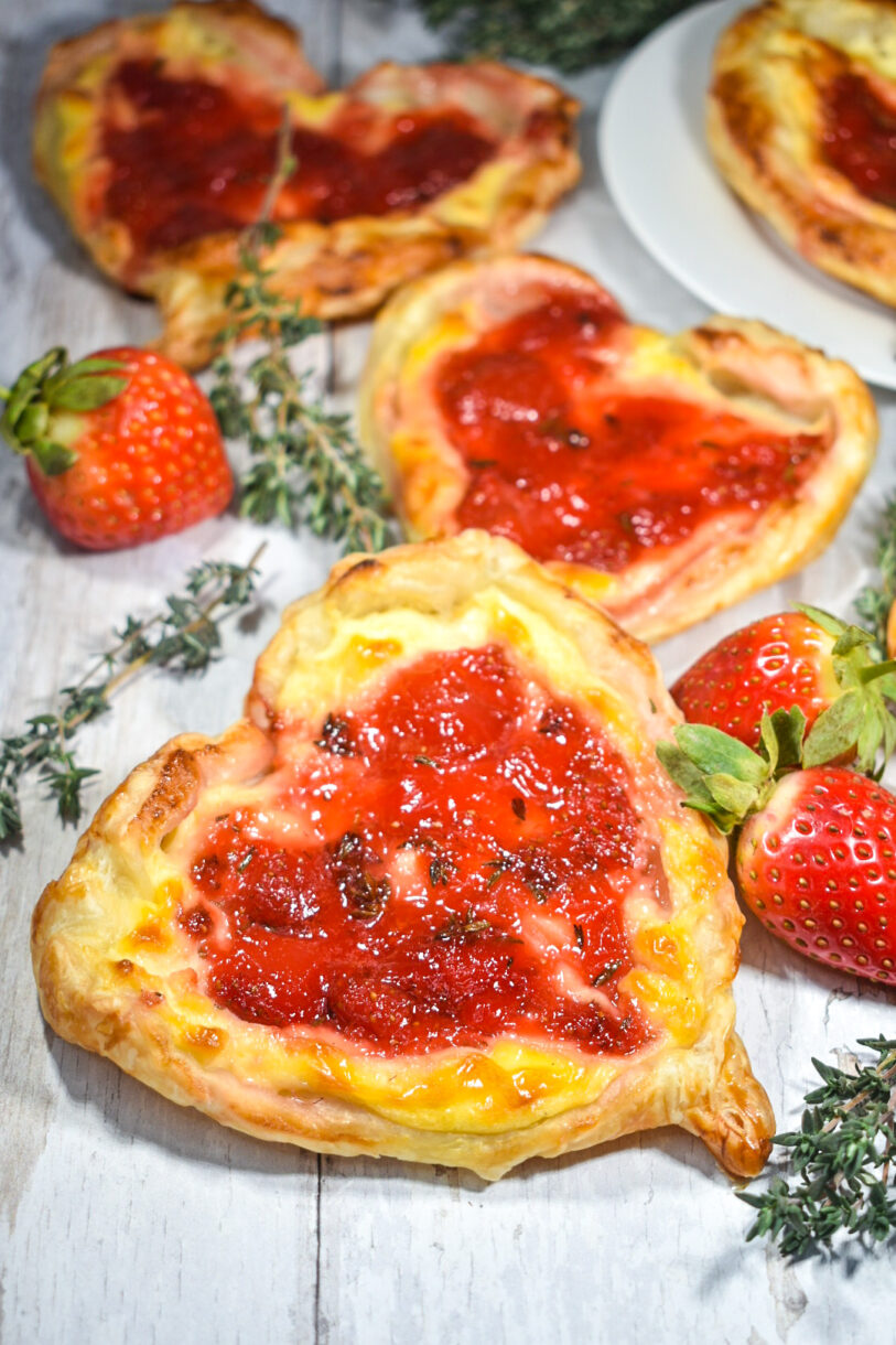 A jam filled puff pastry heart on a white wooden background, with berries and thyme