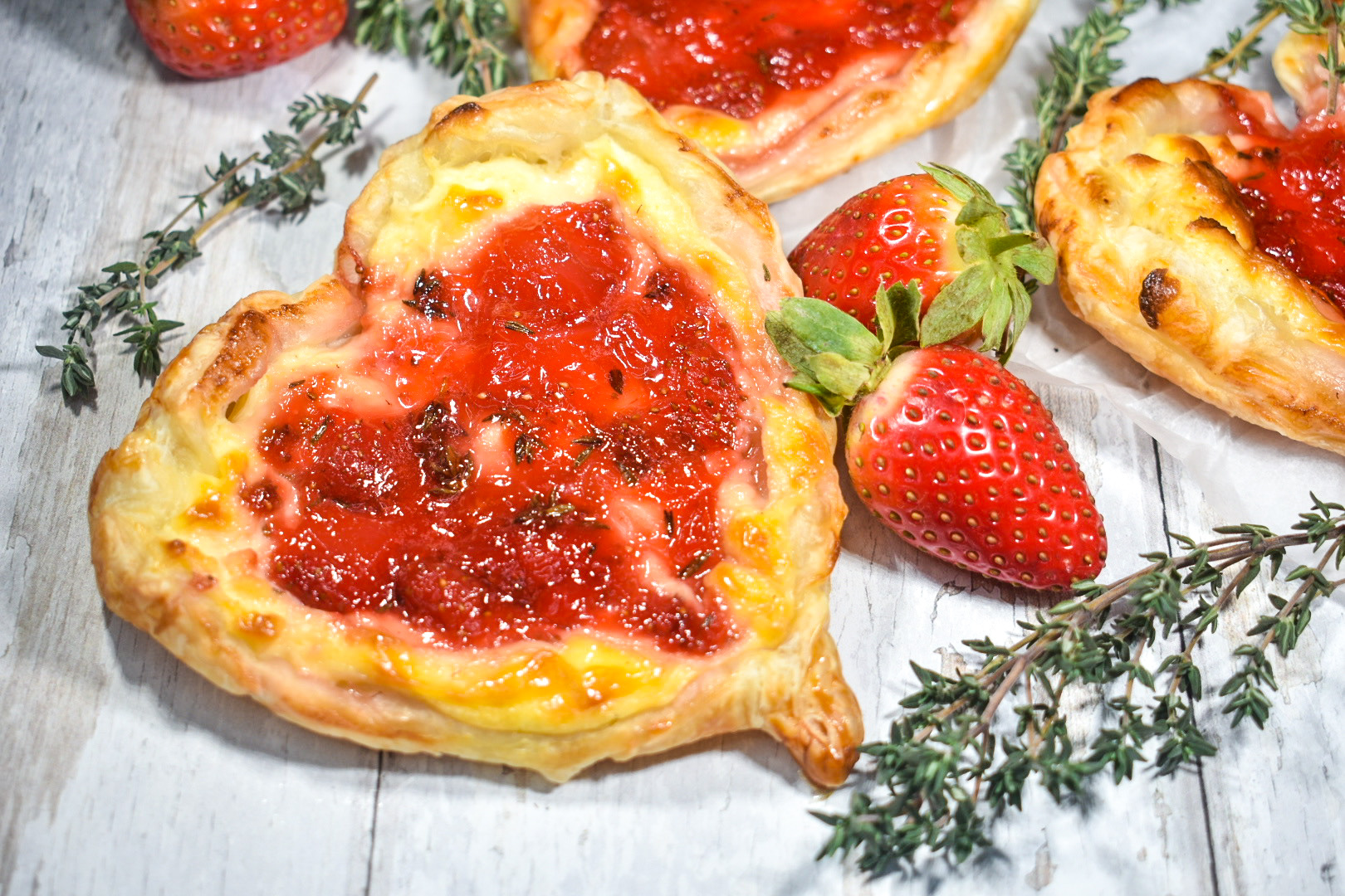 strawberry puff pastry hearts on a white surface with strawberries and a sprig of thyme