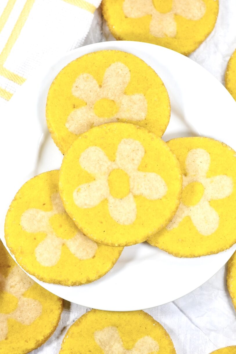 A white plate with vibrant yellow lemon daisy cookies, accompanied by a white and yellow dish cloth, on a white background
