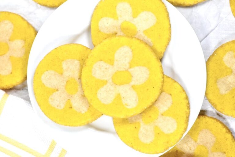 A white plate with vibrant yellow lemon daisy cookies, accompanied by a white and yellow dish cloth, on a white background