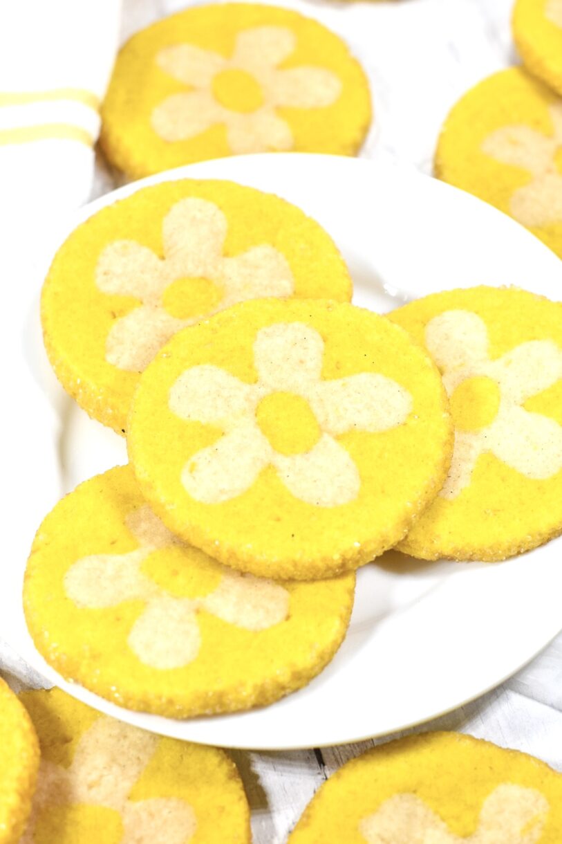 A white plate with vibrant yellow lemon daisy cookies, made from a slice and bake cookie recipe, accompanied by a white and yellow dish cloth, on a white background