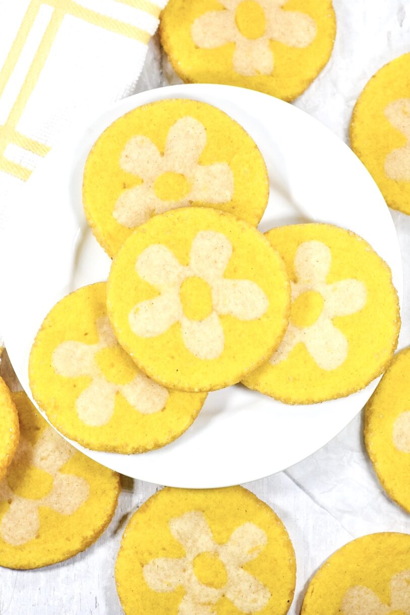A white plate with vibrant yellow lemon daisy cookies, accompanied by a white and yellow dish cloth, on a white background