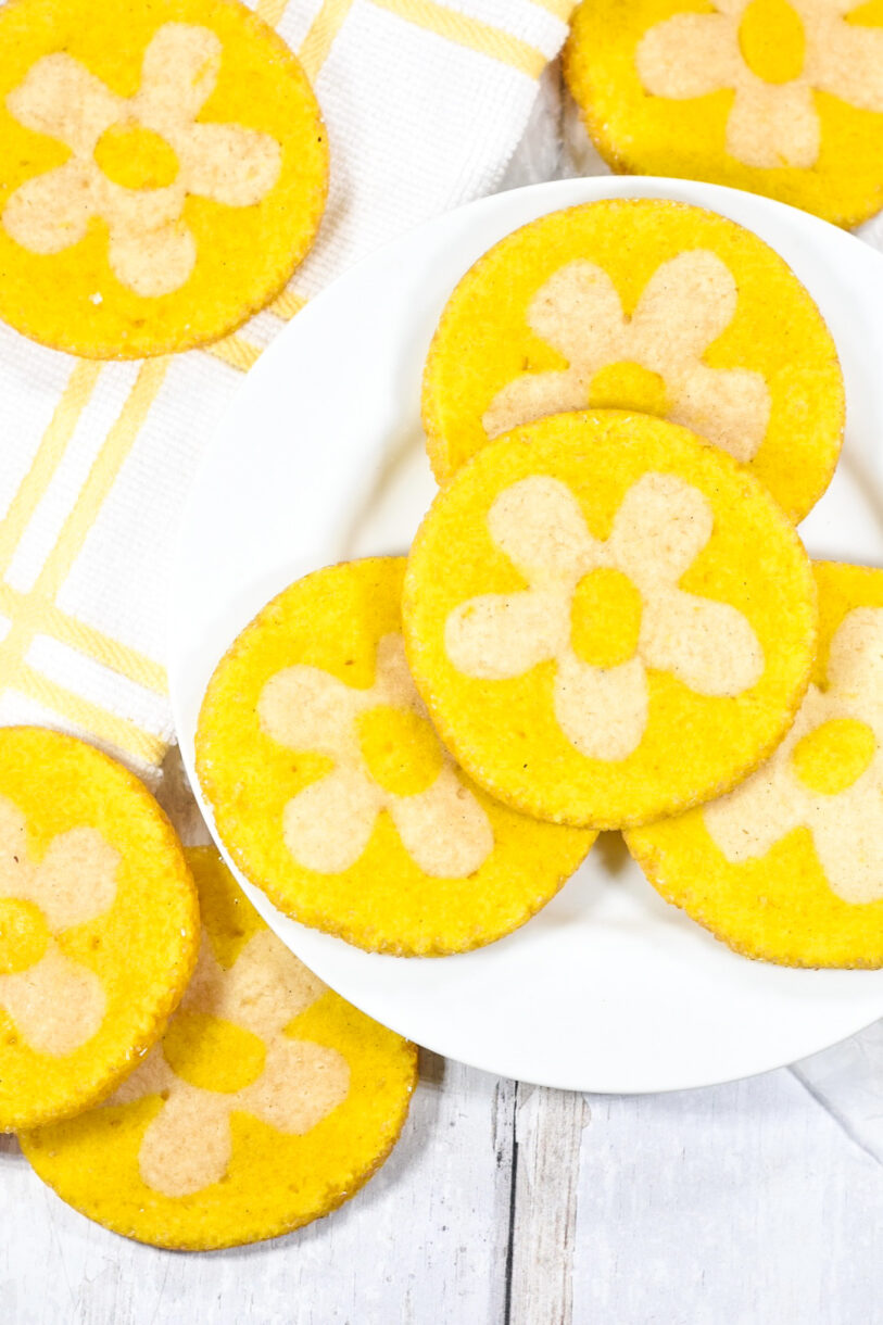 A white plate with vibrant yellow lemon daisy cookies made from a slice and bake cookie recipe, accompanied by a white and yellow dish cloth, on a white background