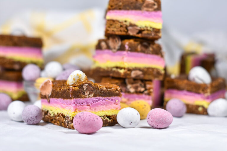 Easter blondies with layers of pastel-colored ganache, surrounded by mini eggs and a yellow and white towel