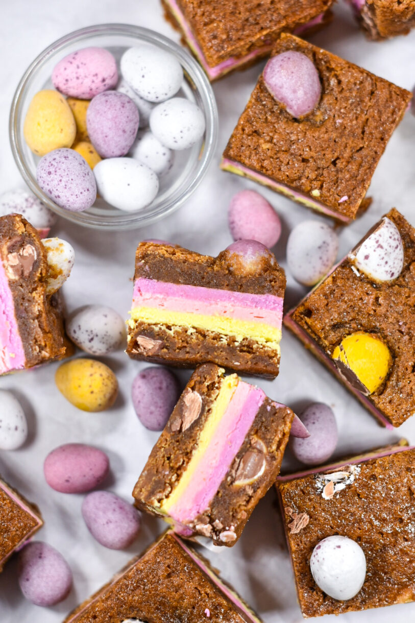 A glass bowl of mini eggs and pastel colored Easter blondies on a white background
