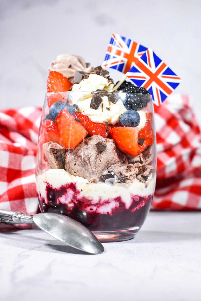 Triple Berry Eton Mess with Chocolate Meringue, layered in a glass with miniature British flags, and red and white gingham cloth in the background