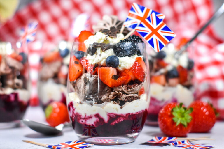 Parfait with chocolate meringue, layered in a glass with miniature British flags, and red and white gingham cloth in the background, and red strawberries