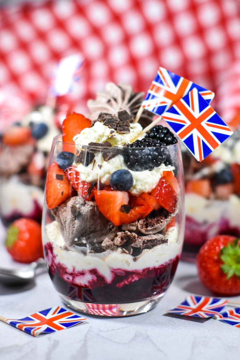 Triple Berry Eton Mess with Chocolate Meringue, layered in a glass with miniature British flags, and red and white gingham cloth in the background