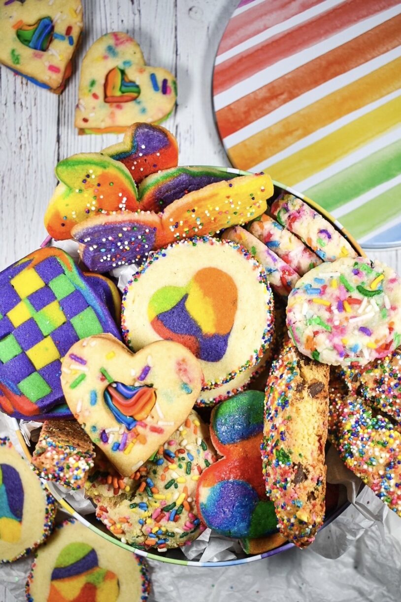 A rainbow striped tin filled with an assortment of rainbow cookies, on a white wooden surface