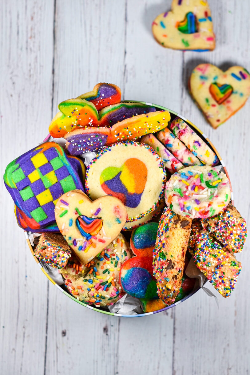 A rainbow striped tin filled with an assortment of rainbow cookies, on a white wooden surface