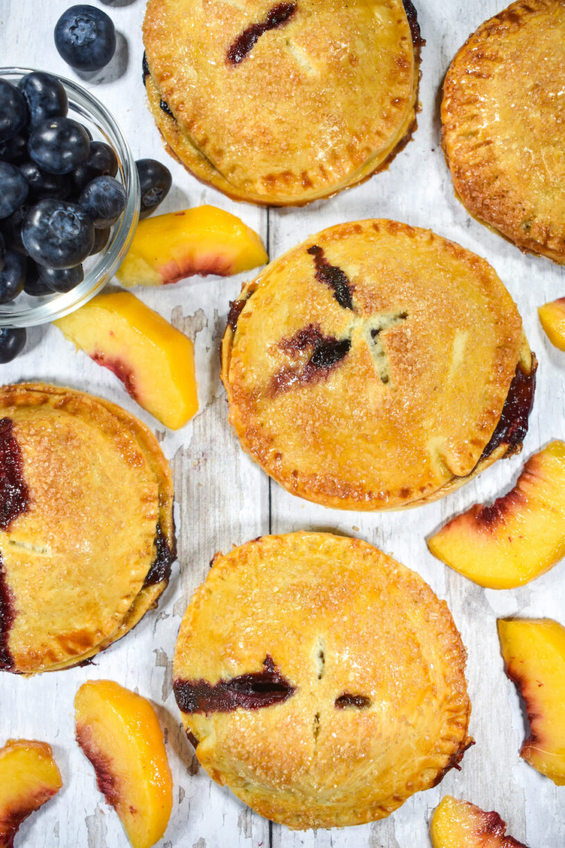 Individually sized blueberry and peach pie arranged on a white surface with fresh fruit