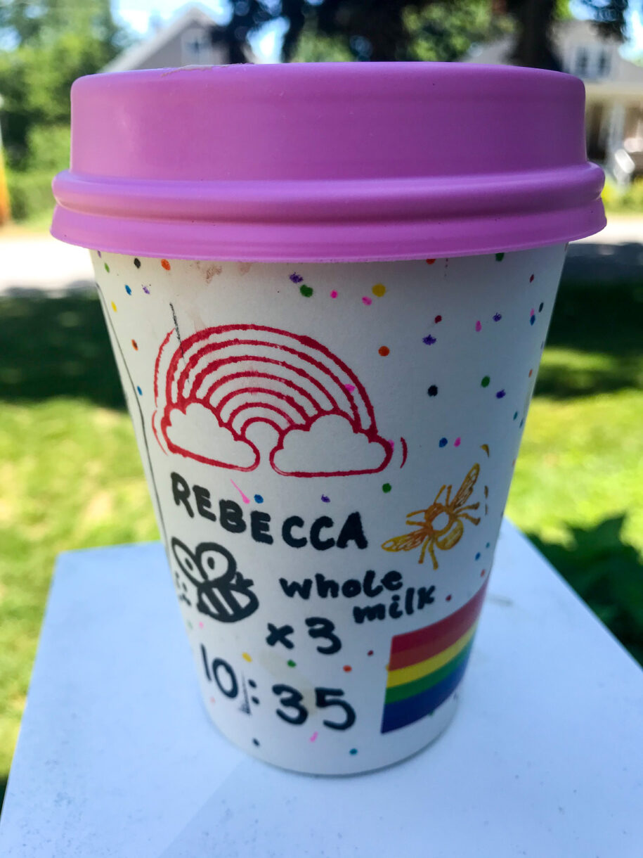 A paper coffee cup with a purple lid, decorated with stamps and rainbow stickers