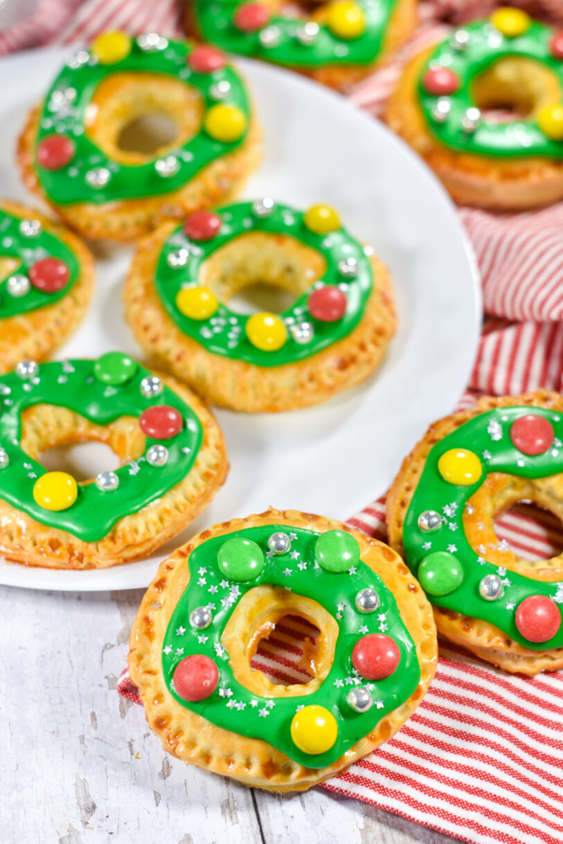 Wreath shaped hand pies with green glaze and candy decorations, and red tea towel, on a white background