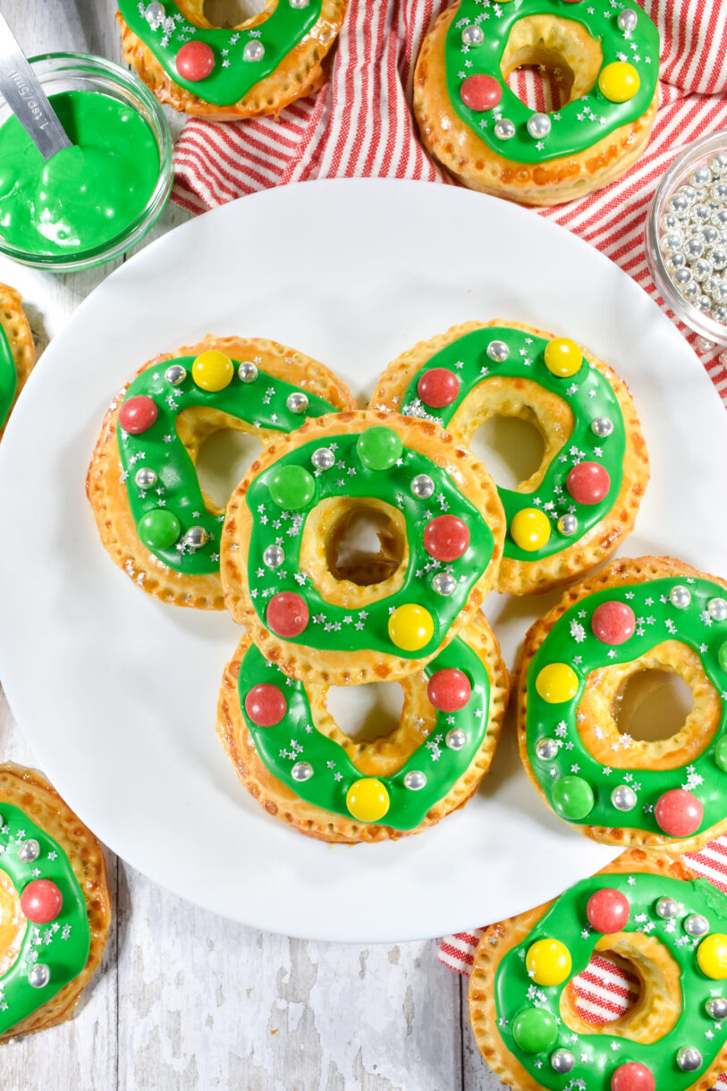 A white plate of wreath shaped pastries with green frosting and candy decorations