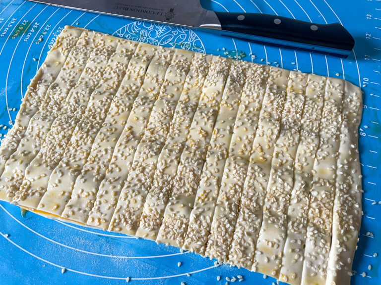 A blue silicone rolling mat with strips of puff pastry coated in sesame seeds, and a chef's knife