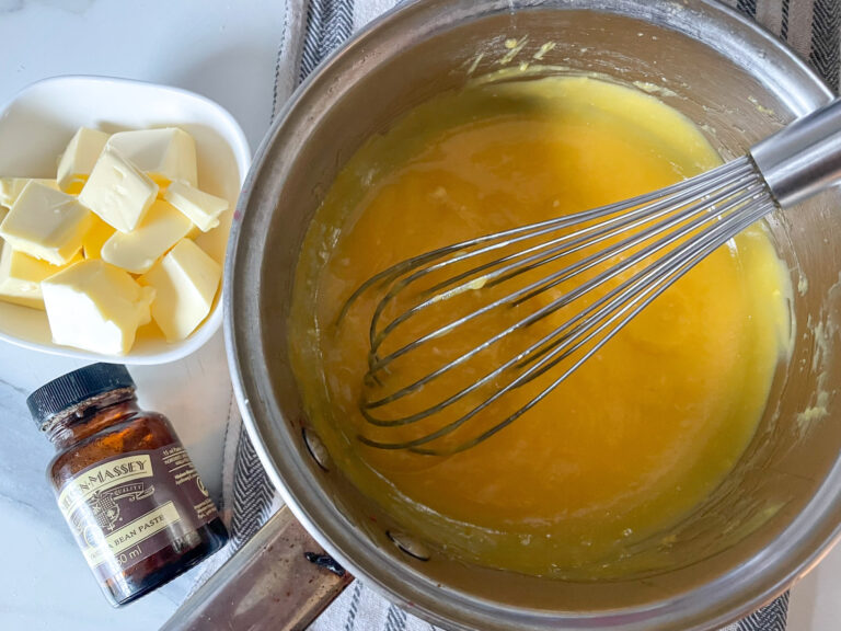 Lemon curd in a saucepan with whisk, alongside cubed butter and a jar of vanilla bean paste