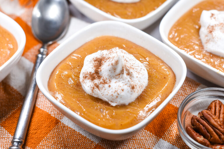 A bowl of butternut squash pudding topped with coconut whipped cream, on an orange checkered towel with a spoon
