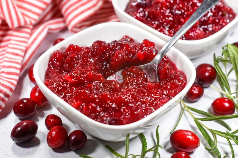 A bowl of cranberry compote surrounded by fresh cranberries and a red and white striped towel