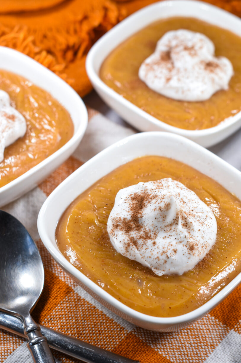 A bowl of butternut squash pudding topped with coconut whipped cream, on an orange checkered towel with a spoon